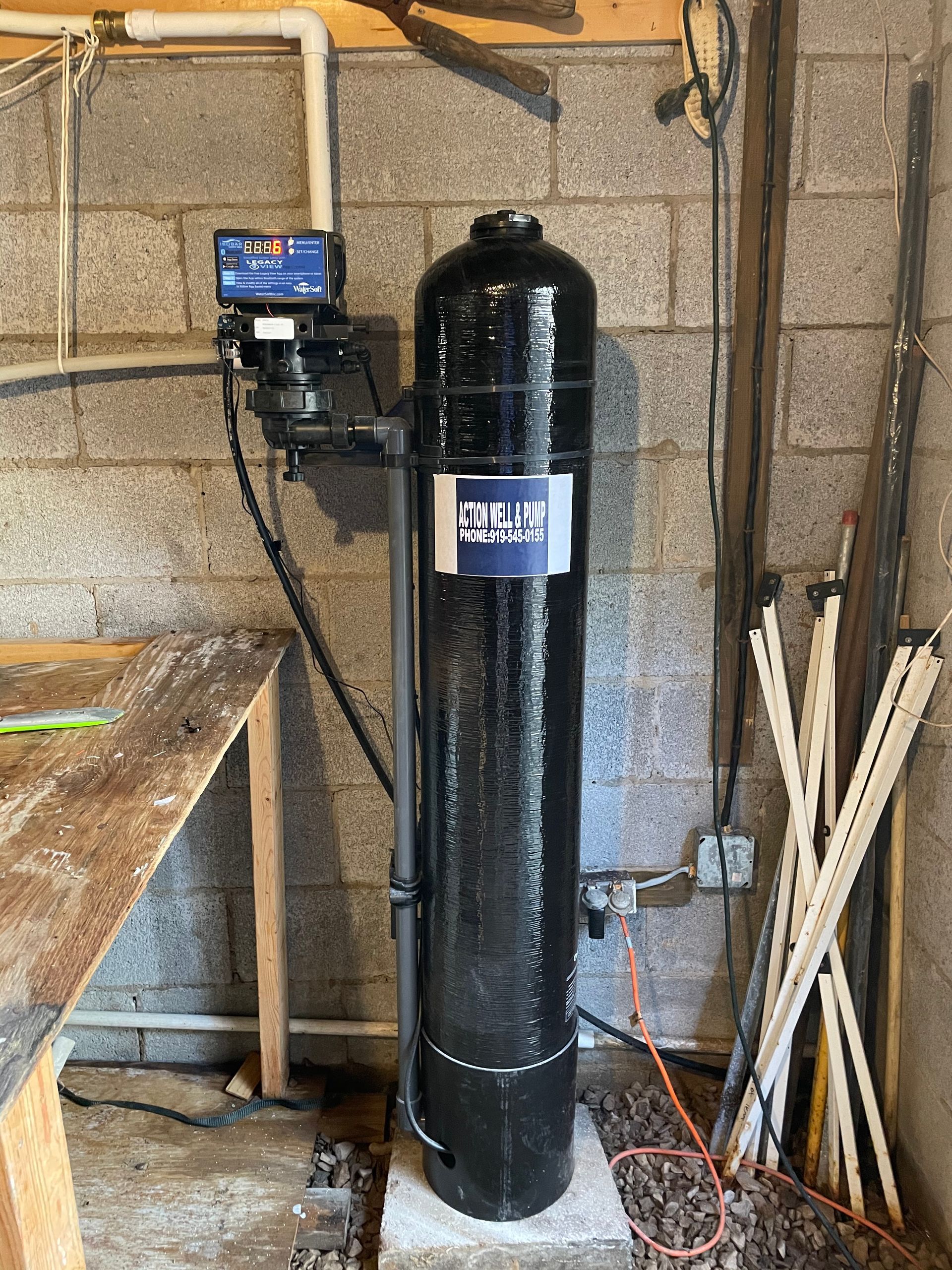 A tall black water filtration tank with a digital control valve stands against a cinder block wall in a utility room.