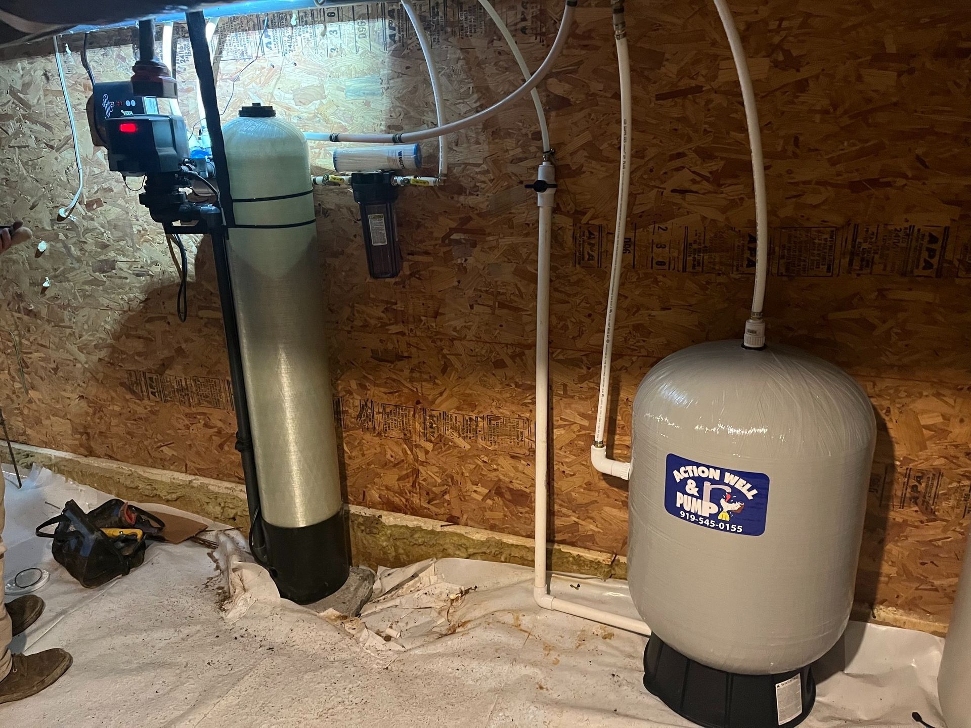 A water treatment system with a tall mineral tank, a small filter, and a grey pressure tank against a wood-paneled wall.