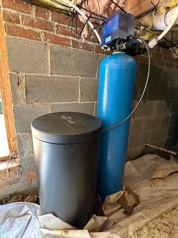 A water softener system with a tall blue mineral tank and a black brine tank installed against a cinder block wall.