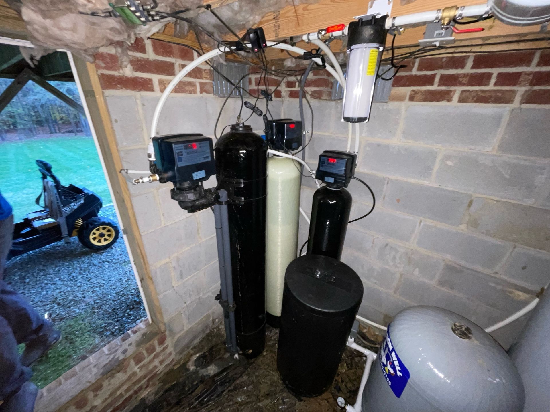 A residential water filtration system with tanks, a clear filter housing, and plumbing, situated against a brick wall.