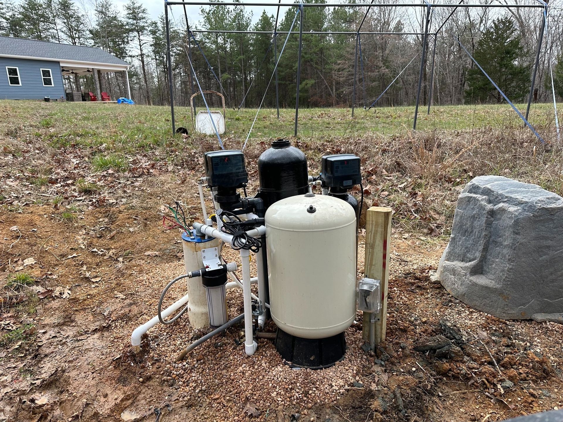 Well pump system with a beige pressure tank and black electrical boxes installed on a lawn in front of a house.