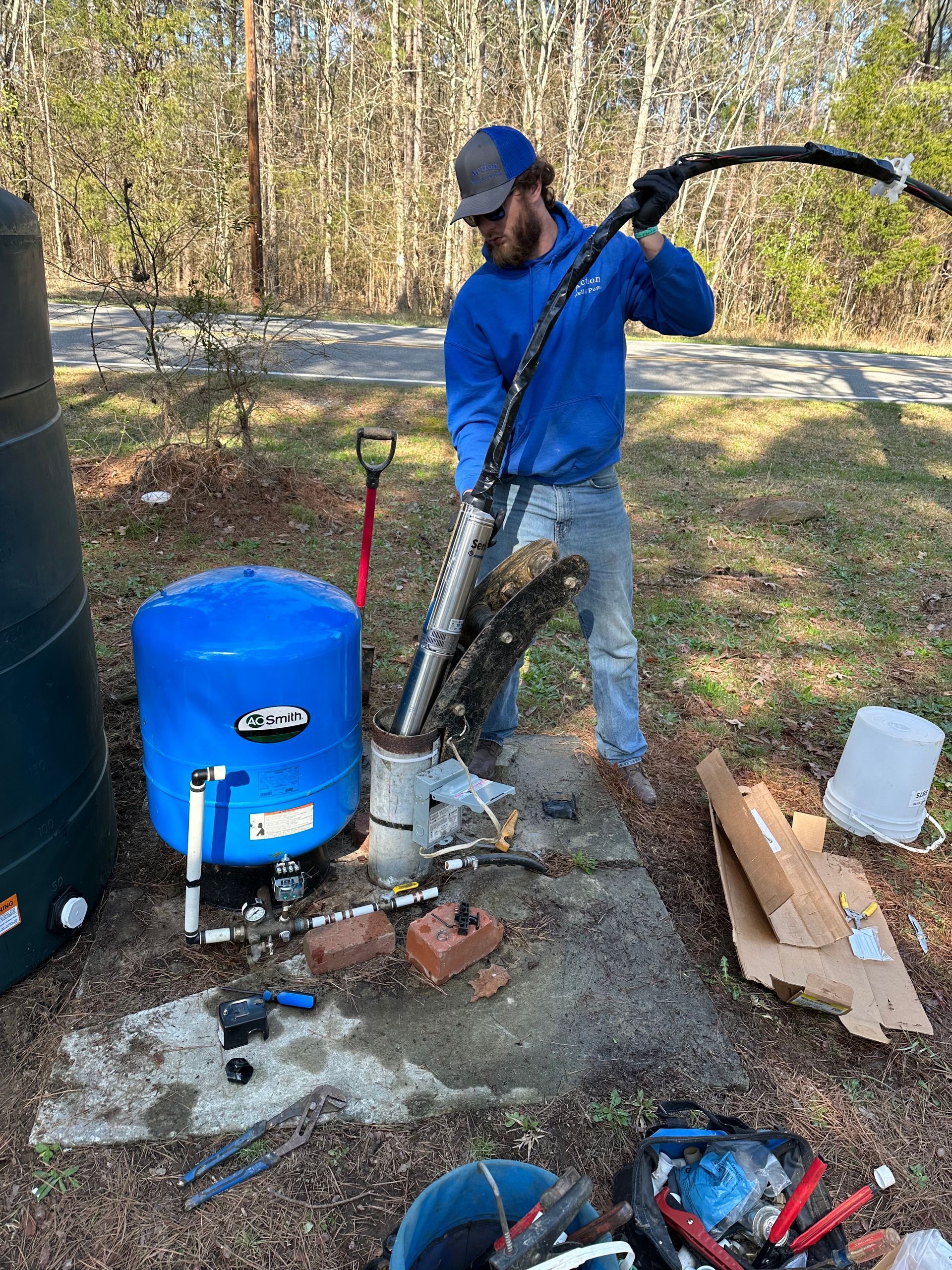 Worker using a vacuum hose beside a blue barrel and tools outdoors in a wooded area