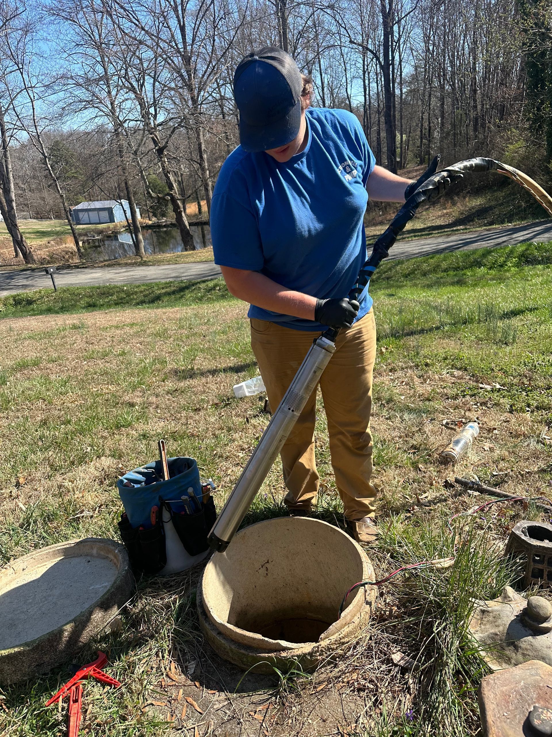 Person in blue shirt using a long tool to work in an open well outdoors.