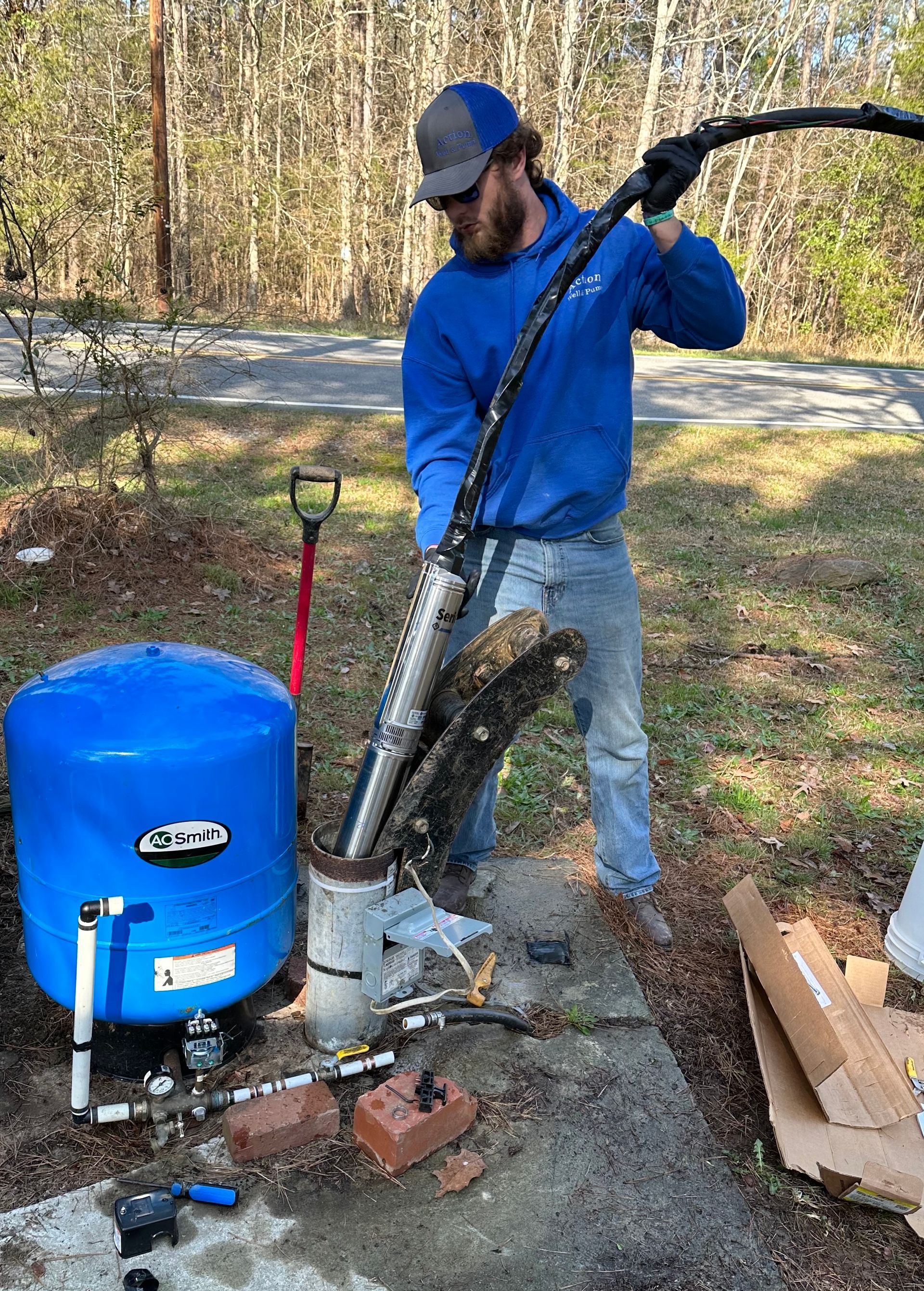 Person operating a leaf vacuum beside a blue collection tank in a wooded yard