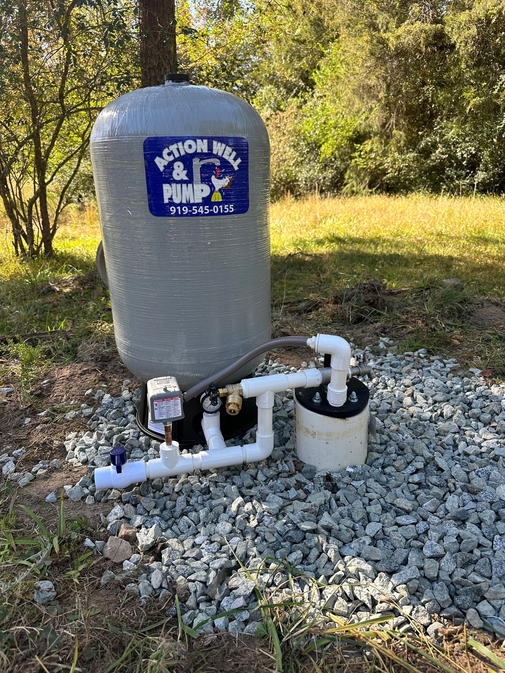 Grey utility tank and white pipes on gravel beside a wooded area, with a small blue sign on the tank.