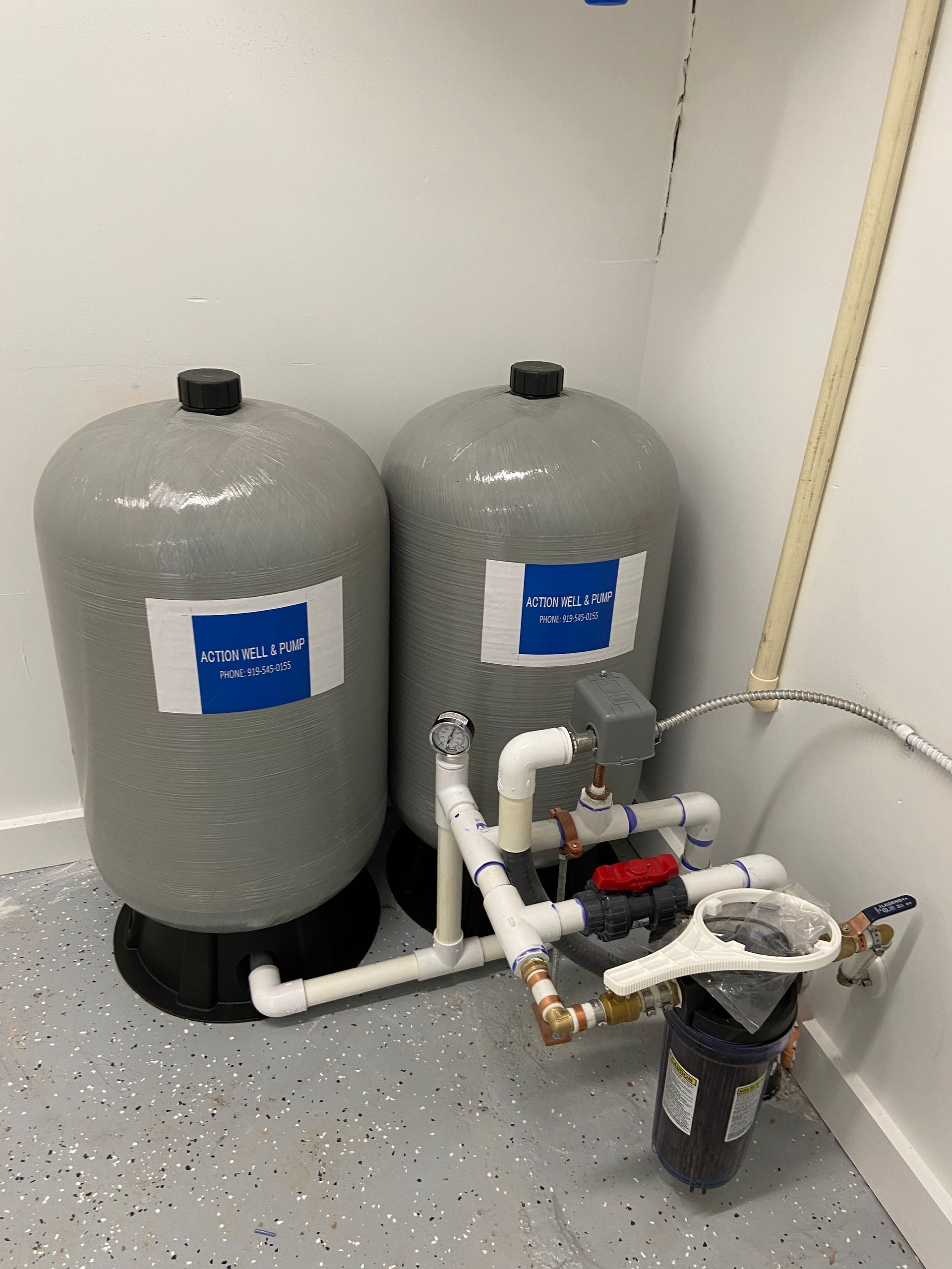 Two gray water softener tanks with plumbing and a control valve in a utility room corner