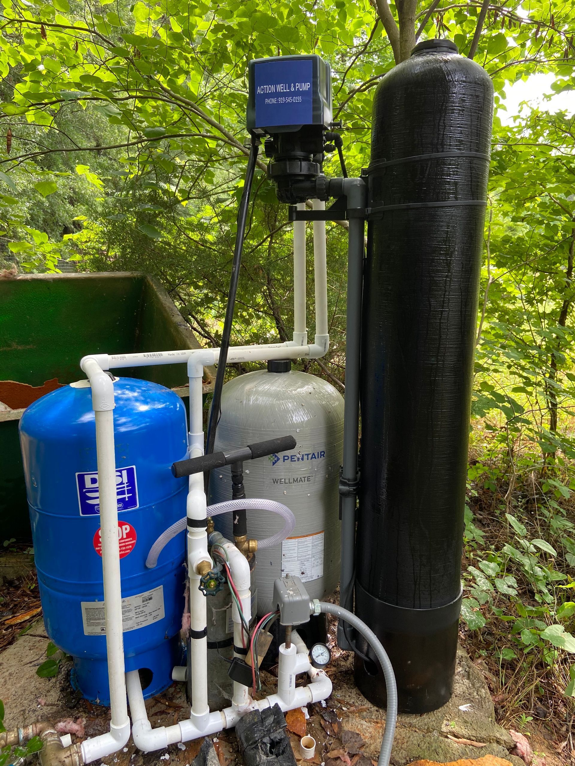 Outdoor water filtration system with blue tank, white pipes, gray tank, and black insulated cylinder beside a green wall