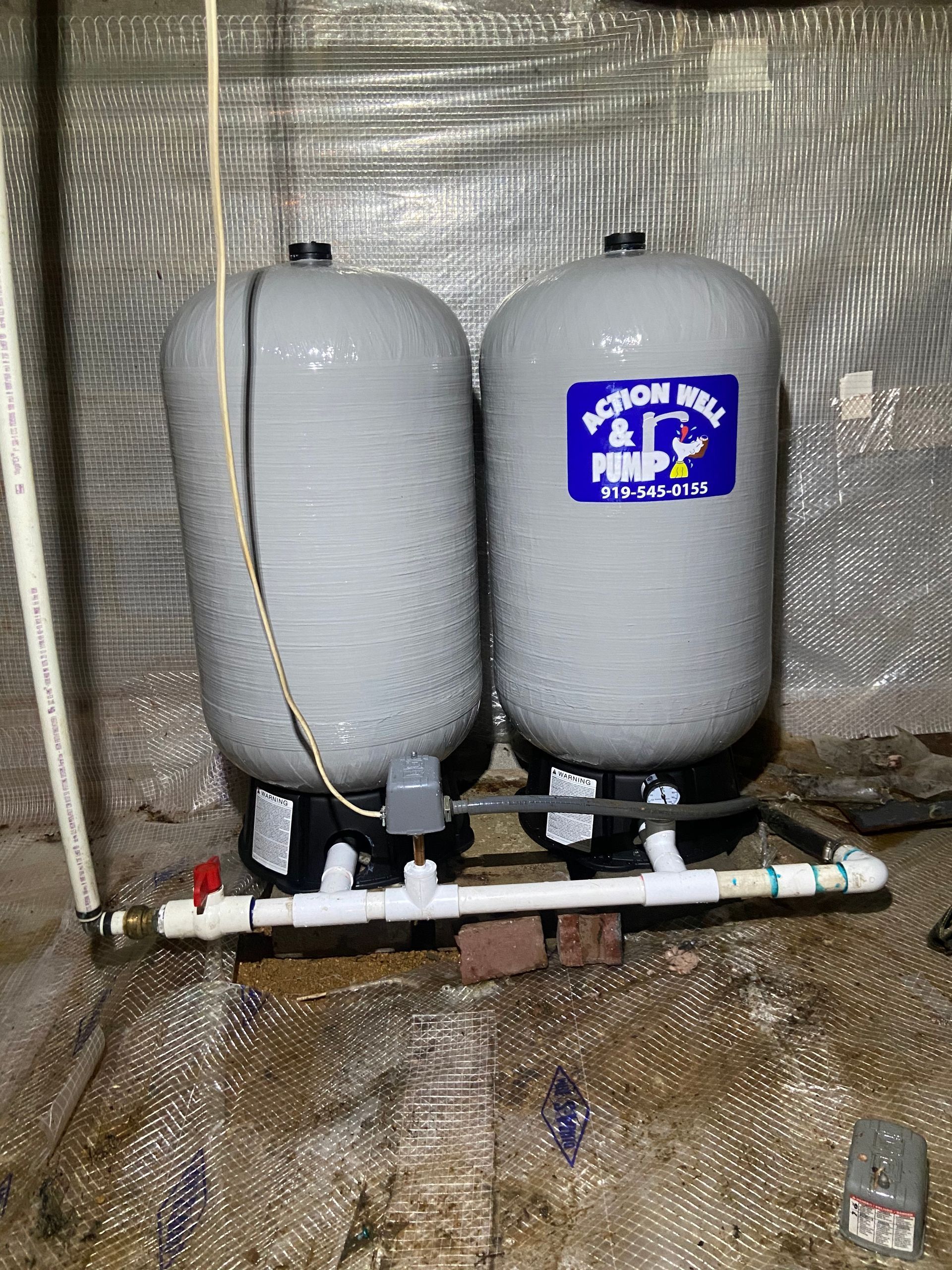 Two gray water tanks connected to white piping in a utility area with reflective insulation behind them.