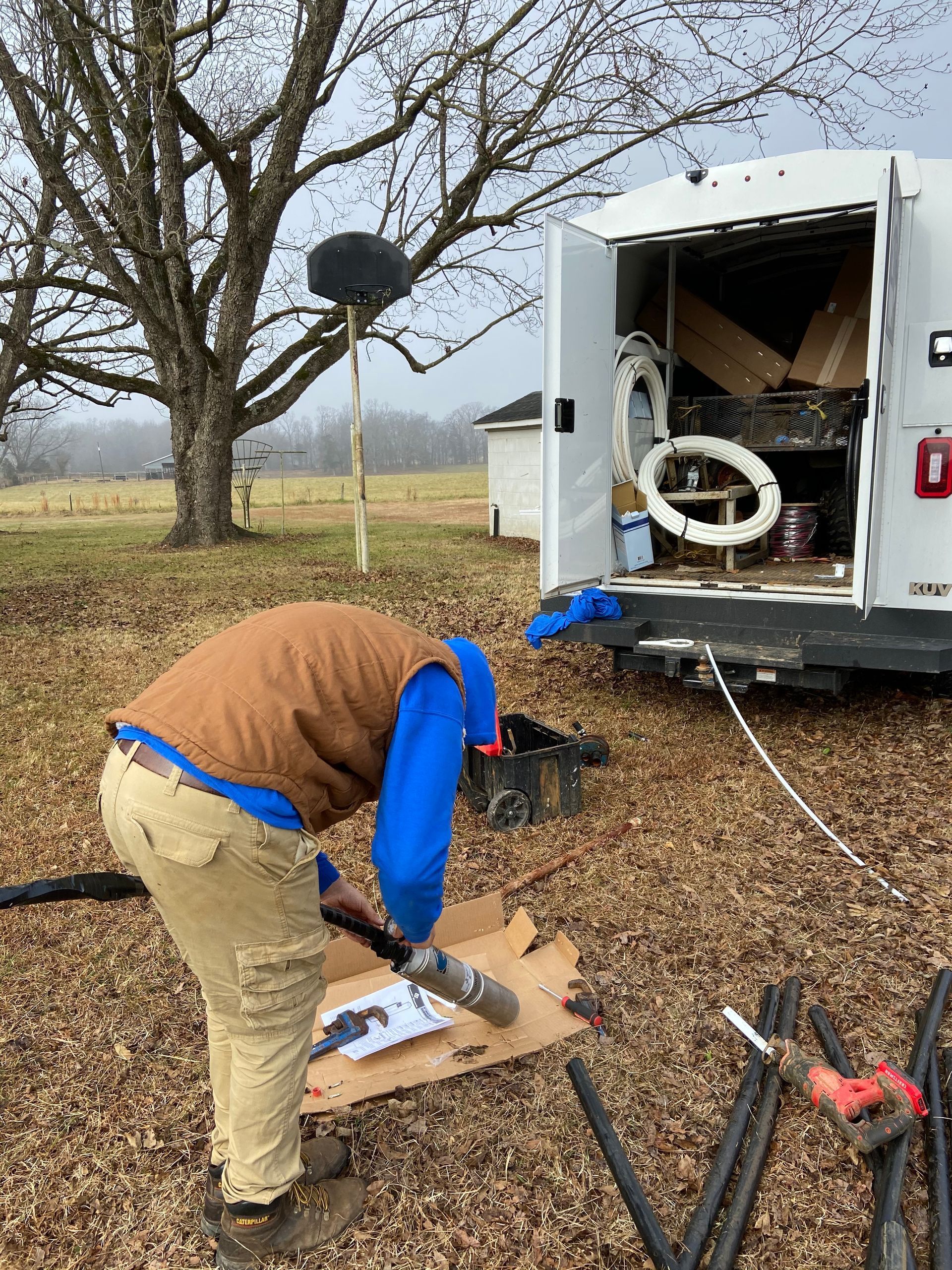 Person working beside open utility trailer on a grassy field, handling tools and pipes.