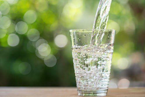 Clear water pouring into a textured glass against a blurred, sunlit green garden background.
