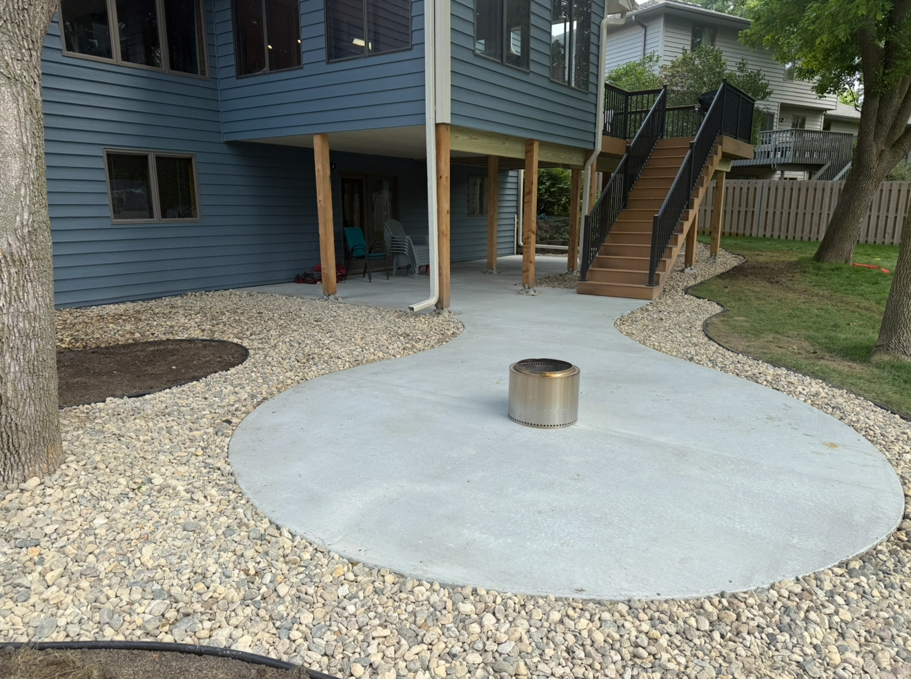 Concrete patio with fire pit, surrounded by gravel, connected to a blue house with stairs.