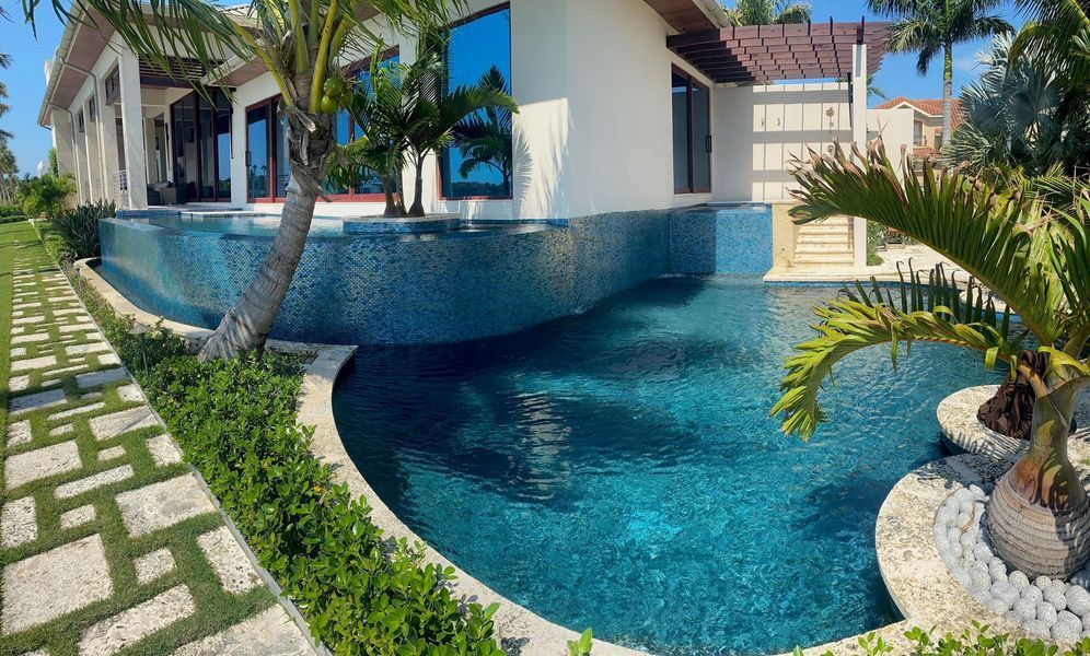 Luxurious house with a blue-tiled infinity pool. Lush greenery, palm trees, and stone walkway surround it.