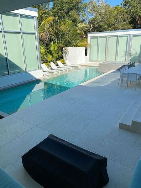 Modern outdoor space with a pool, white deck, lounge chairs, and buildings with large windows, surrounded by trees.