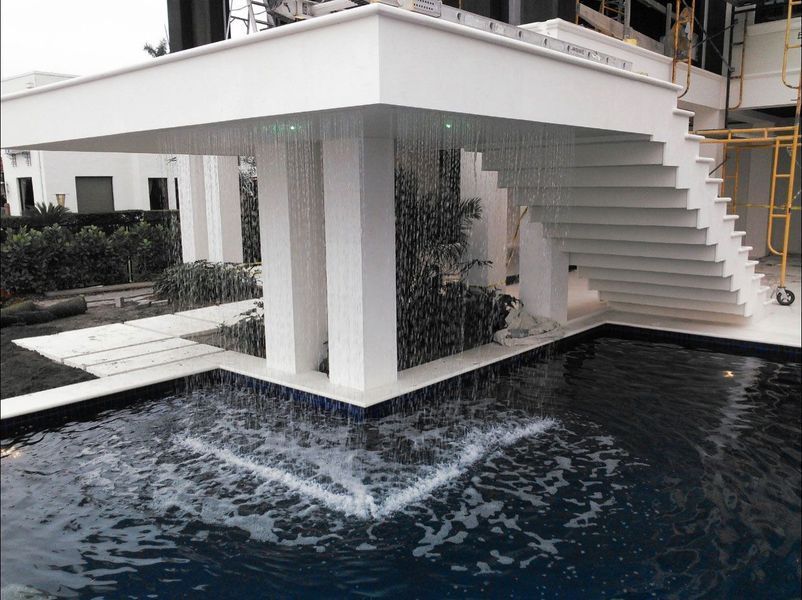White structure with waterfall feature cascading into a dark blue pool. Stairs and greenery are also visible.