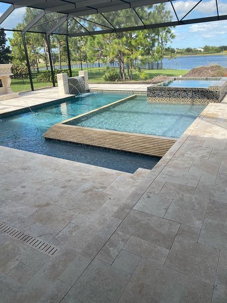 Swimming pool with a bridge feature, surrounded by tan patio and screened enclosure, overlooking a lake.