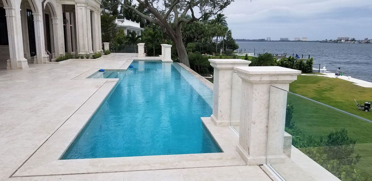 A luxurious pool, beside a building with columns, overlooking a waterfront on a cloudy day.