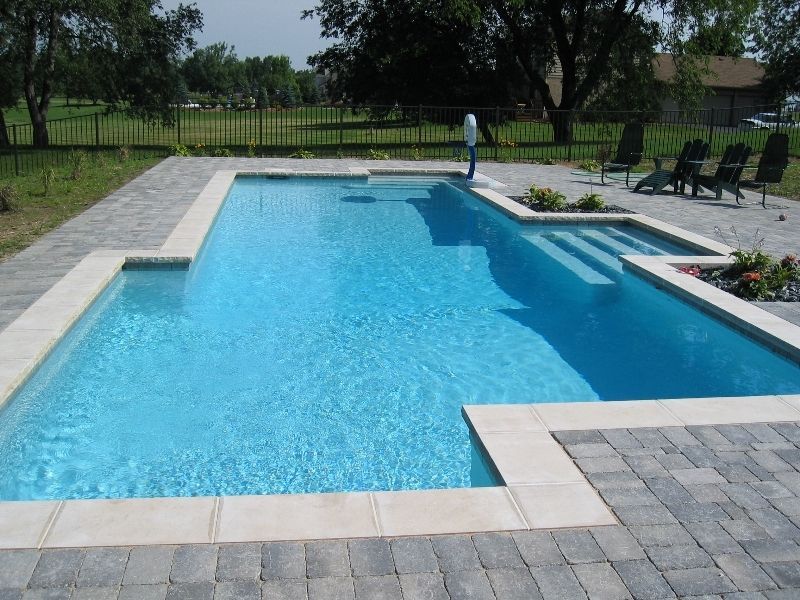Rectangular swimming pool with light blue water and stone patio. Lounge chairs sit nearby.
