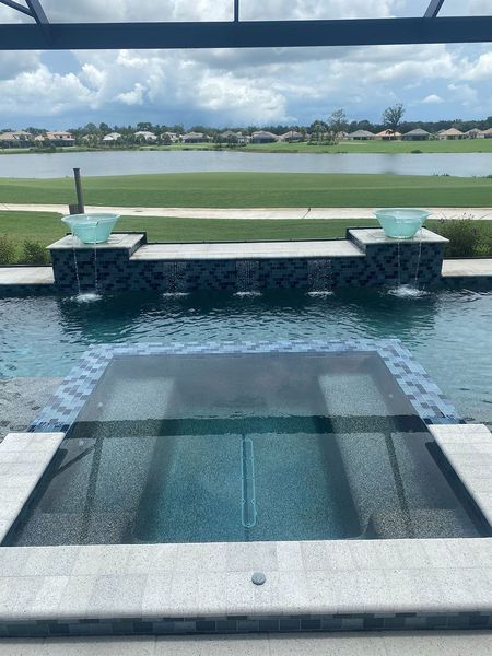 Pool with fountains and a spa, overlooking a golf course and lake under a cloudy sky.