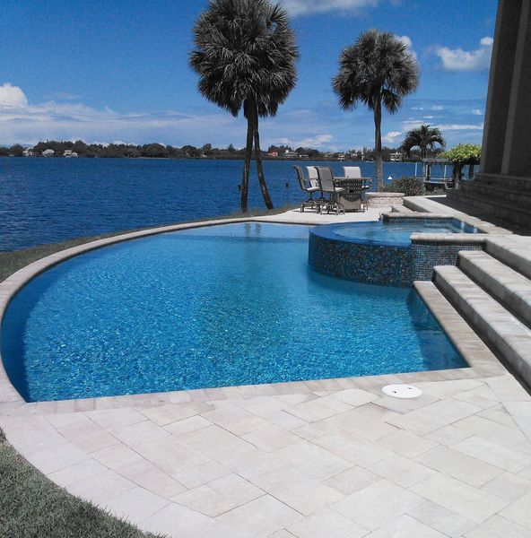 Blue pool and spa with water view, palm trees, and stone patio.