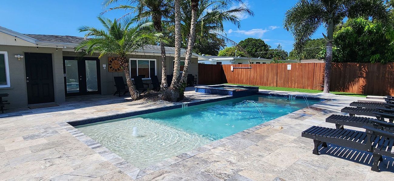 A backyard with a pool, lounge chairs, a fence, and a house under a clear, blue sky.