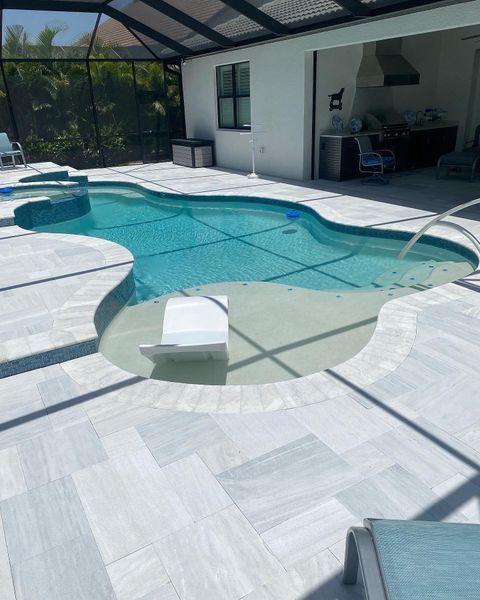 Pool with attached spa, surrounded by light-colored tile, next to a covered patio area.