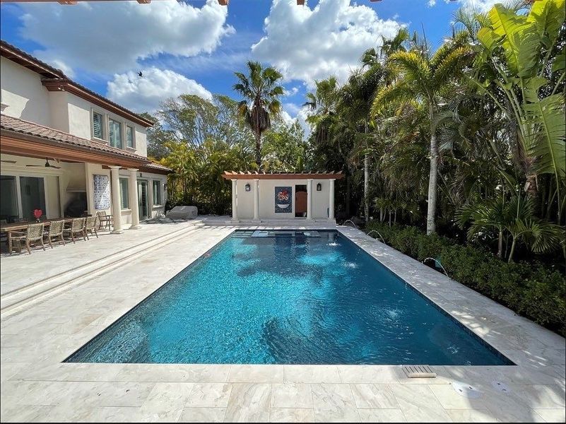 Swimming pool and cabana next to a house with palm trees under a blue sky.