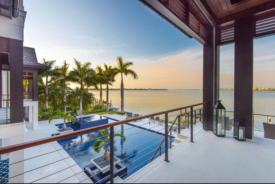 Luxury balcony overlooking a pool and ocean at sunset, framed by palm trees and dark wood pillars.