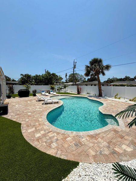 Backyard pool with tan pavers, blue water, lounge chairs, and palm tree under a sunny sky.
