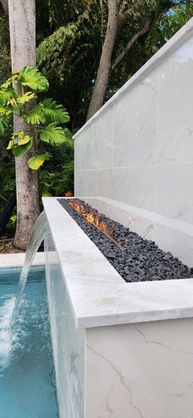 Water flows from a white stone wall into a pool with a linear fire feature filled with black rocks.