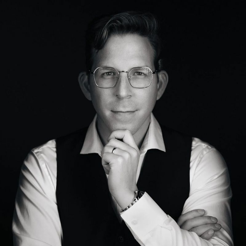 Man in glasses, holding chin, wearing a vest and collared shirt, black and white portrait.