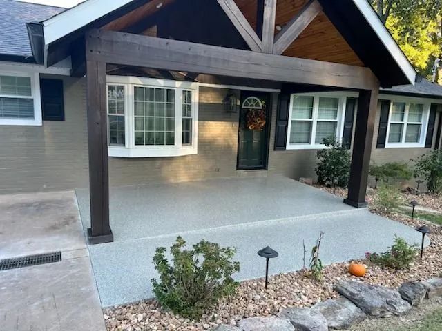 Front porch with dark wood beams, light gray siding, white windows, and a stone walkway with shrubs.
