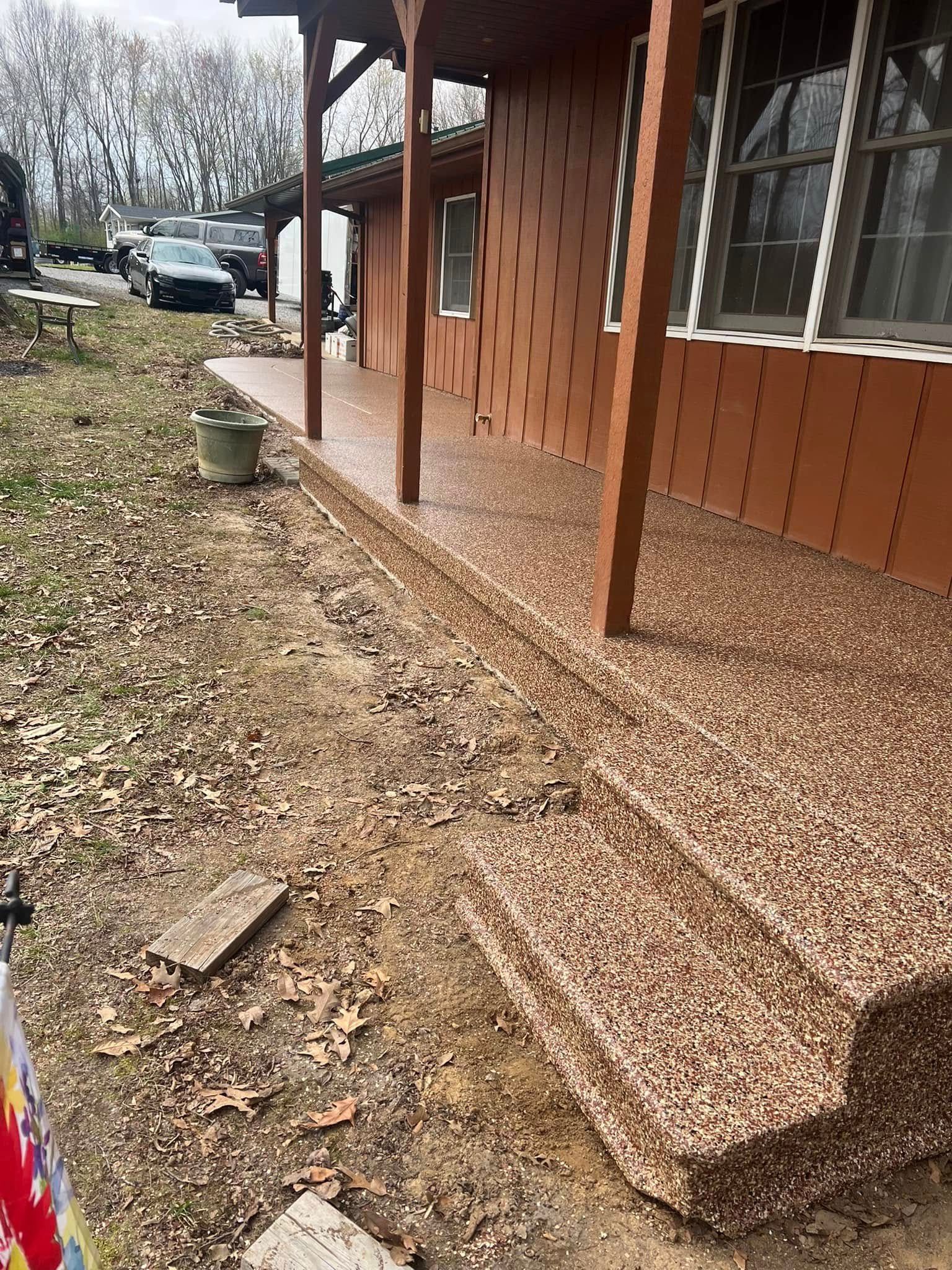 Front porch of a brown house with wood posts, gravel ground, and a yard with parked cars in the background