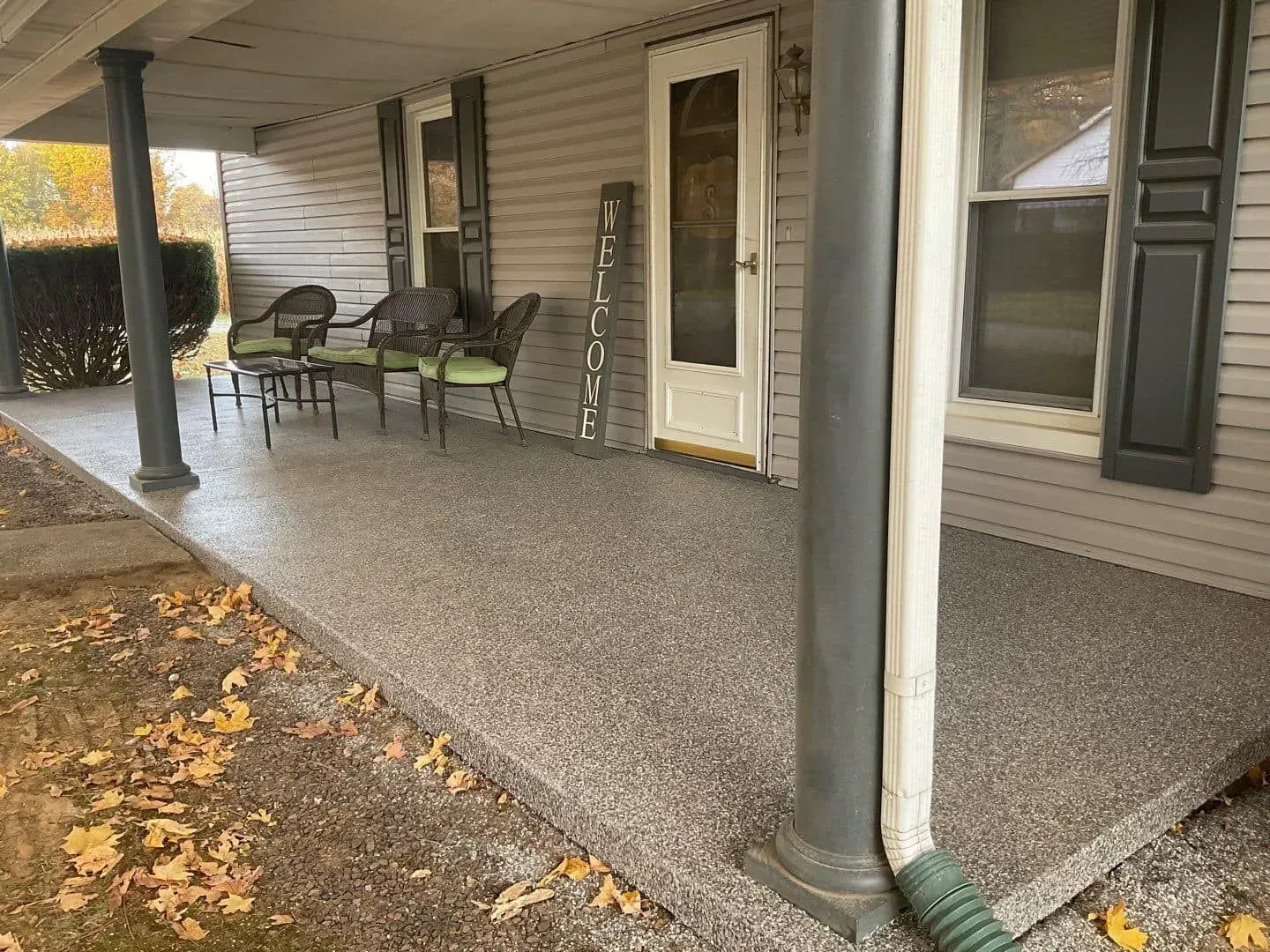 Covered porch with chairs, white door, and wet pebbled floor beside a house in autumn.