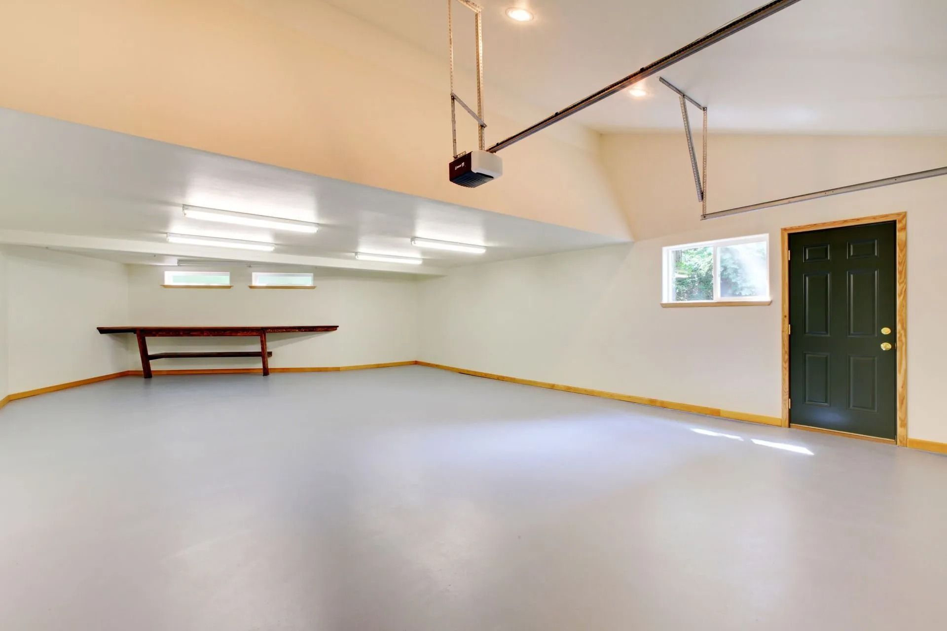 Empty garage with white walls, gray floor, overhead lights, workbench, and a green door.
