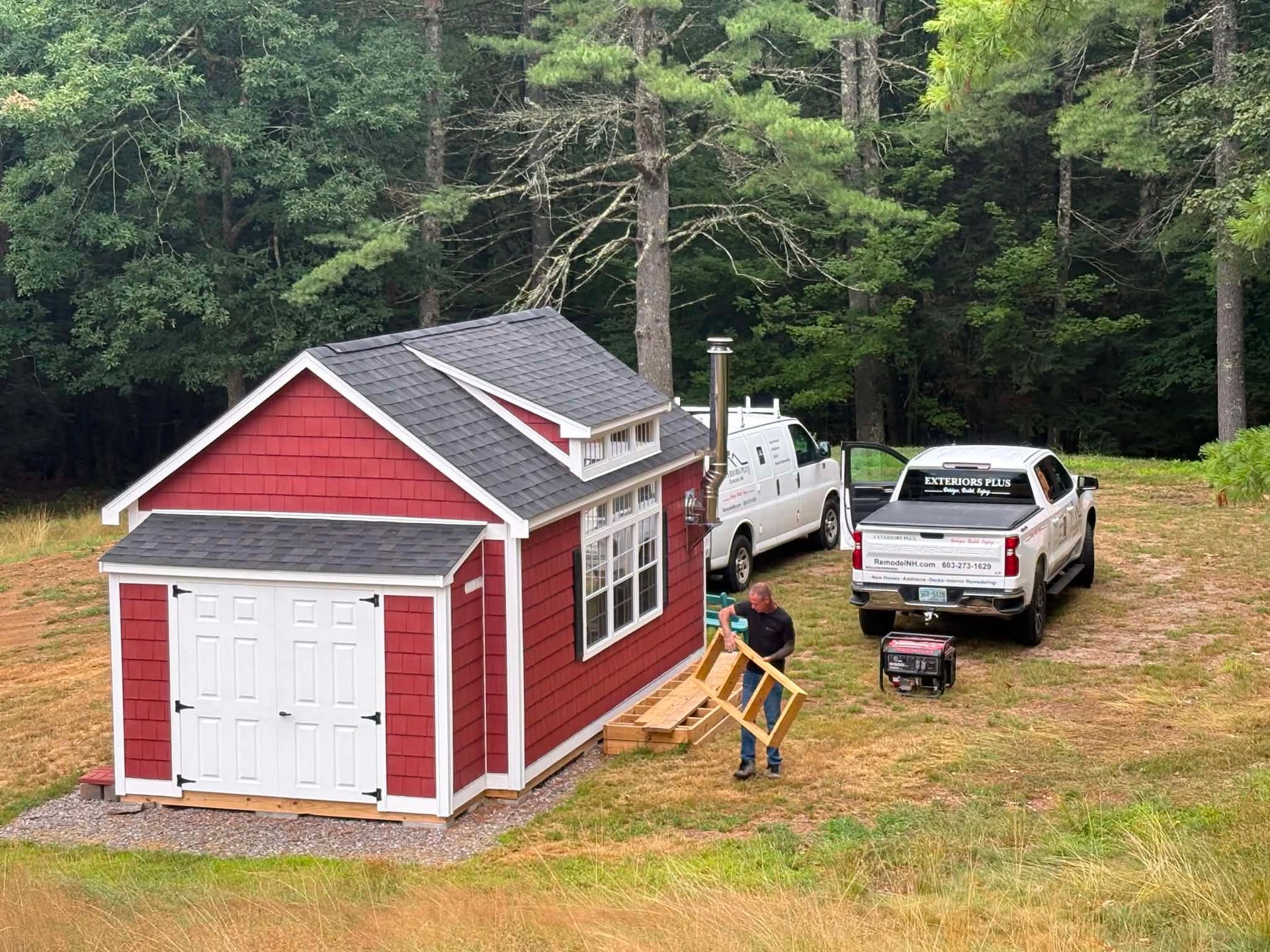 Red shed-style tiny house in a wooded yard, with two white pickup trucks and tools nearby.