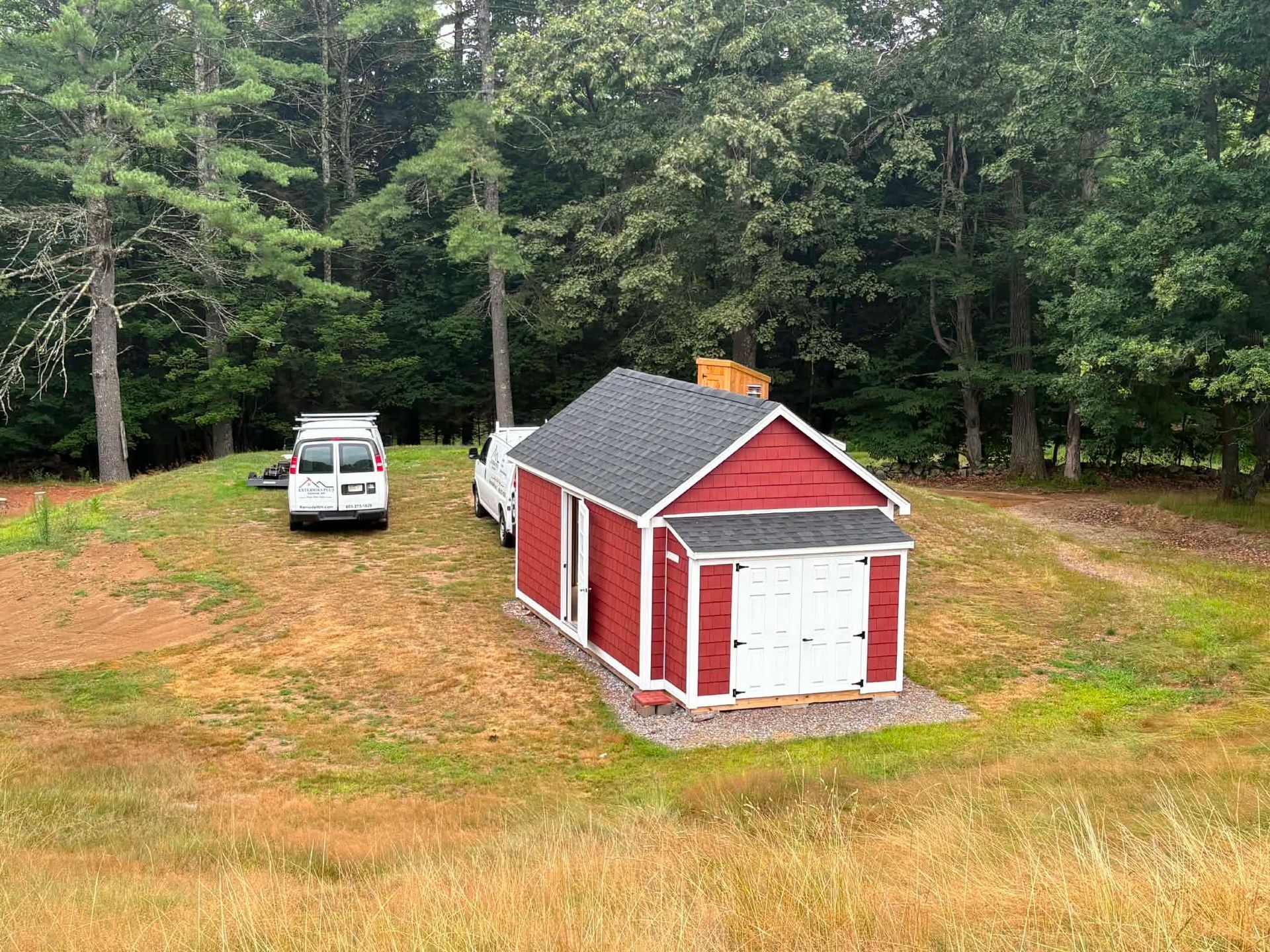 Red shed with white doors beside a white van in a grassy clearing near dense woods