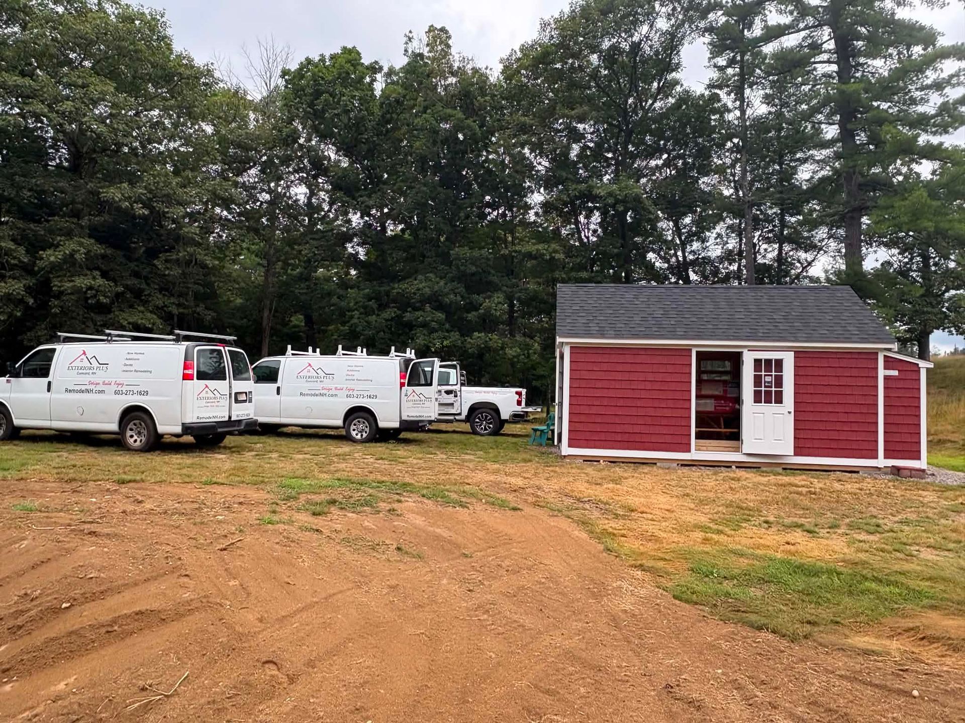 Red shed-style building with white trim beside white service vans on a dirt lot, backed by trees.