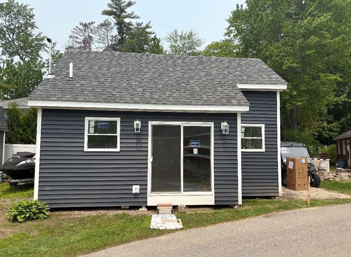 Small dark-blue house with white-trimmed sliding glass door and gray shingled roof in a wooded setting
