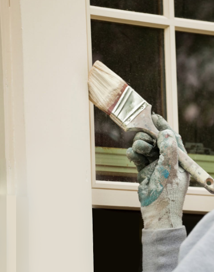 Hand wearing a paint-splattered glove painting a cream window frame with a brush