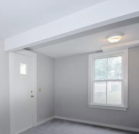 Small empty gray room with a white door, window, carpet, and ceiling light.