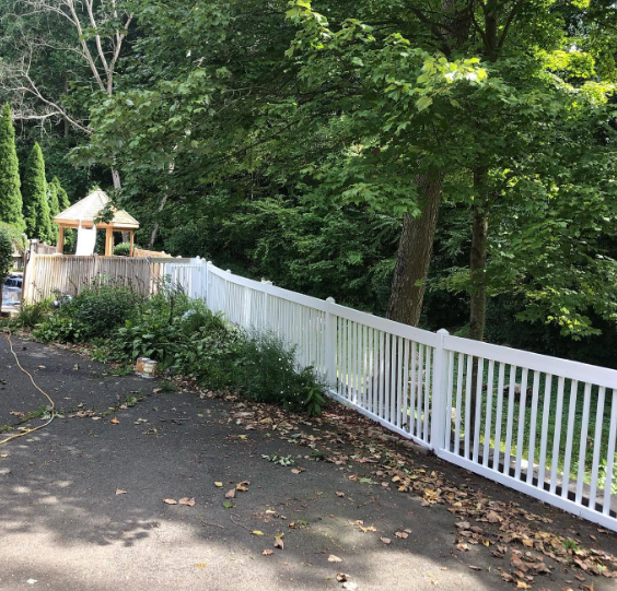 White fence along a shaded driveway beside dense green trees and a small gazebo