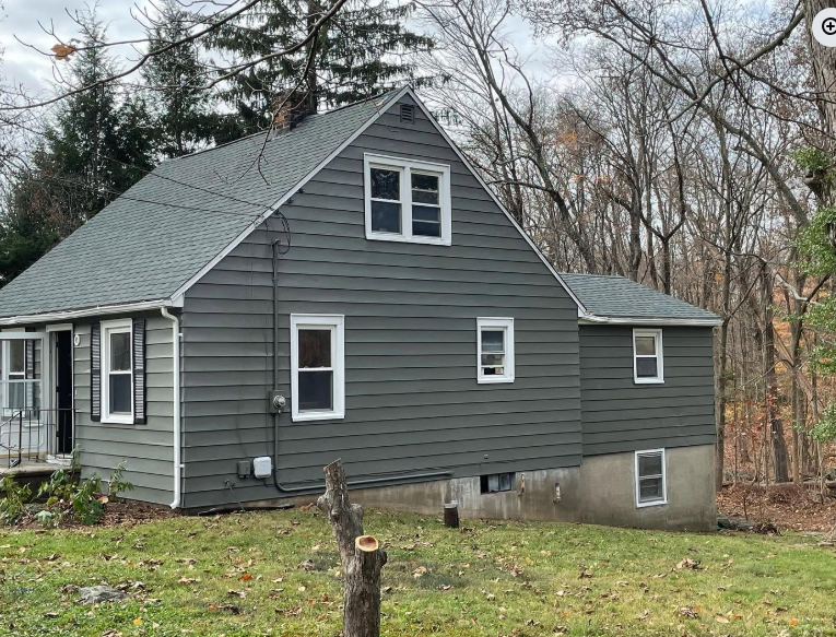Gray house with steep roof and porch, set in a wooded yard on an overcast day