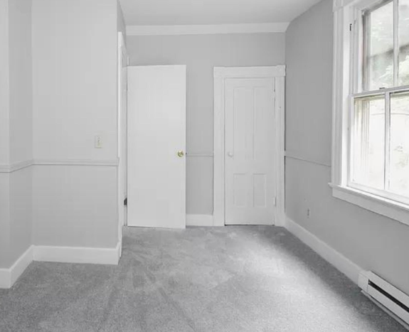 Empty gray room with carpet, white trim, open door, and window light on the right.