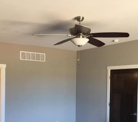 Bedroom corner with gray walls, white vent, dark door, and a ceiling fan with light.
