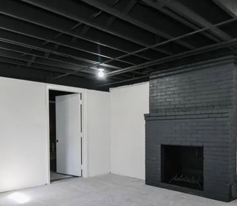 Empty room with white walls, exposed black ceiling beams, and a dark brick fireplace.