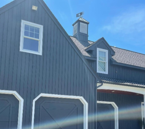 Dark blue barn-style house with white trim, dormer windows, and a garage under a bright blue sky