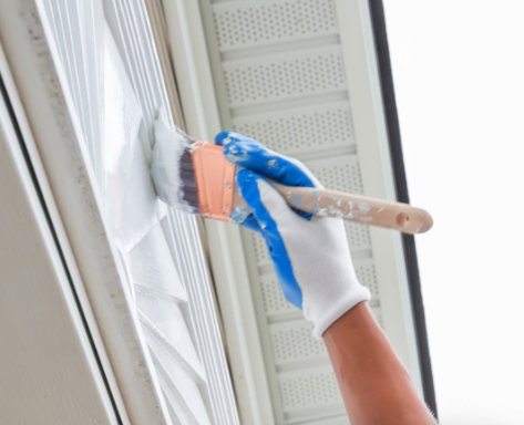Gloved hand painting white trim with a brush near a window