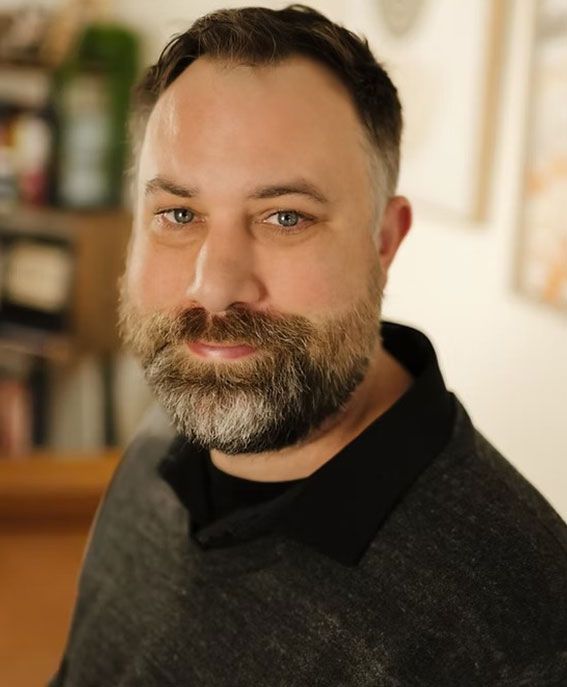 Portrait of a bearded man in a dark sweater, standing indoors with a soft, blurred background