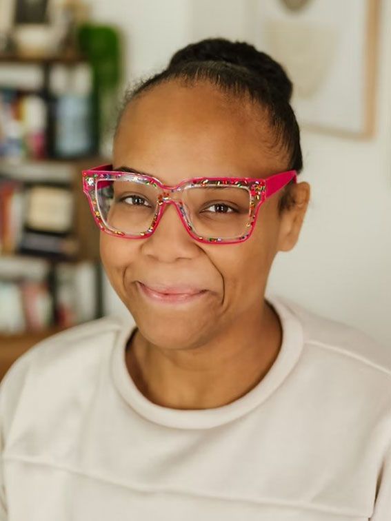 Smiling person wearing red glasses and a cream top against a softly blurred indoor background