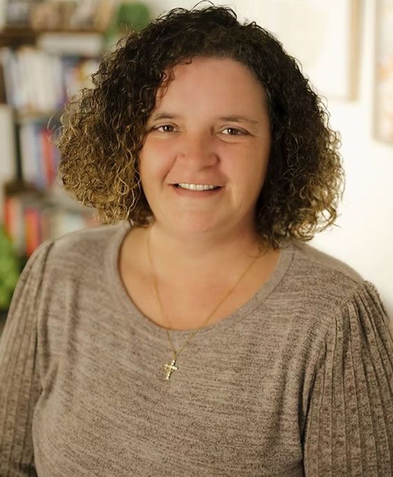 Smiling person with curly hair wearing a beige top and gold cross necklace in a softly lit indoor setting